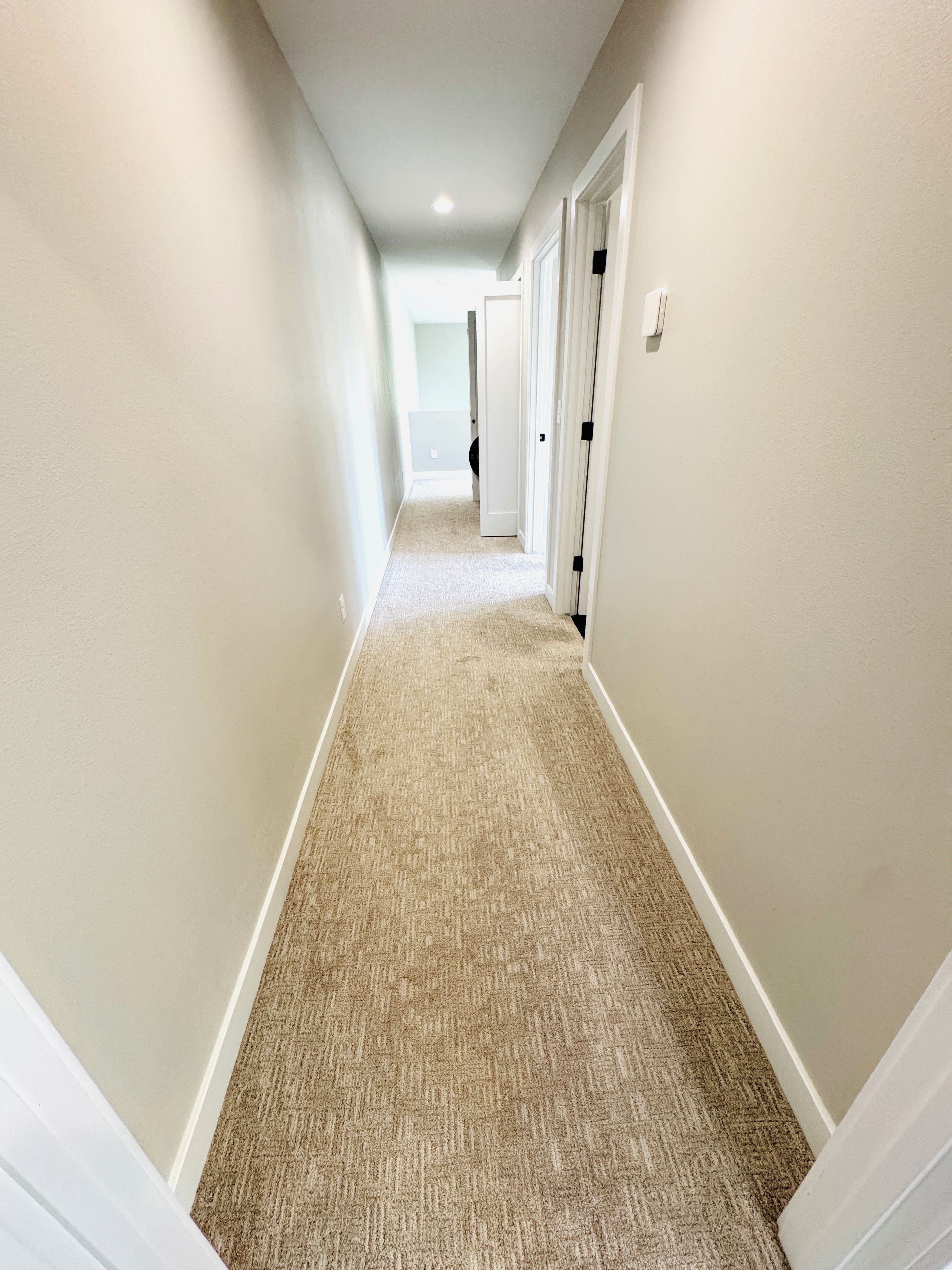 A carpeted hallway with white walls and doors, leading to another room.