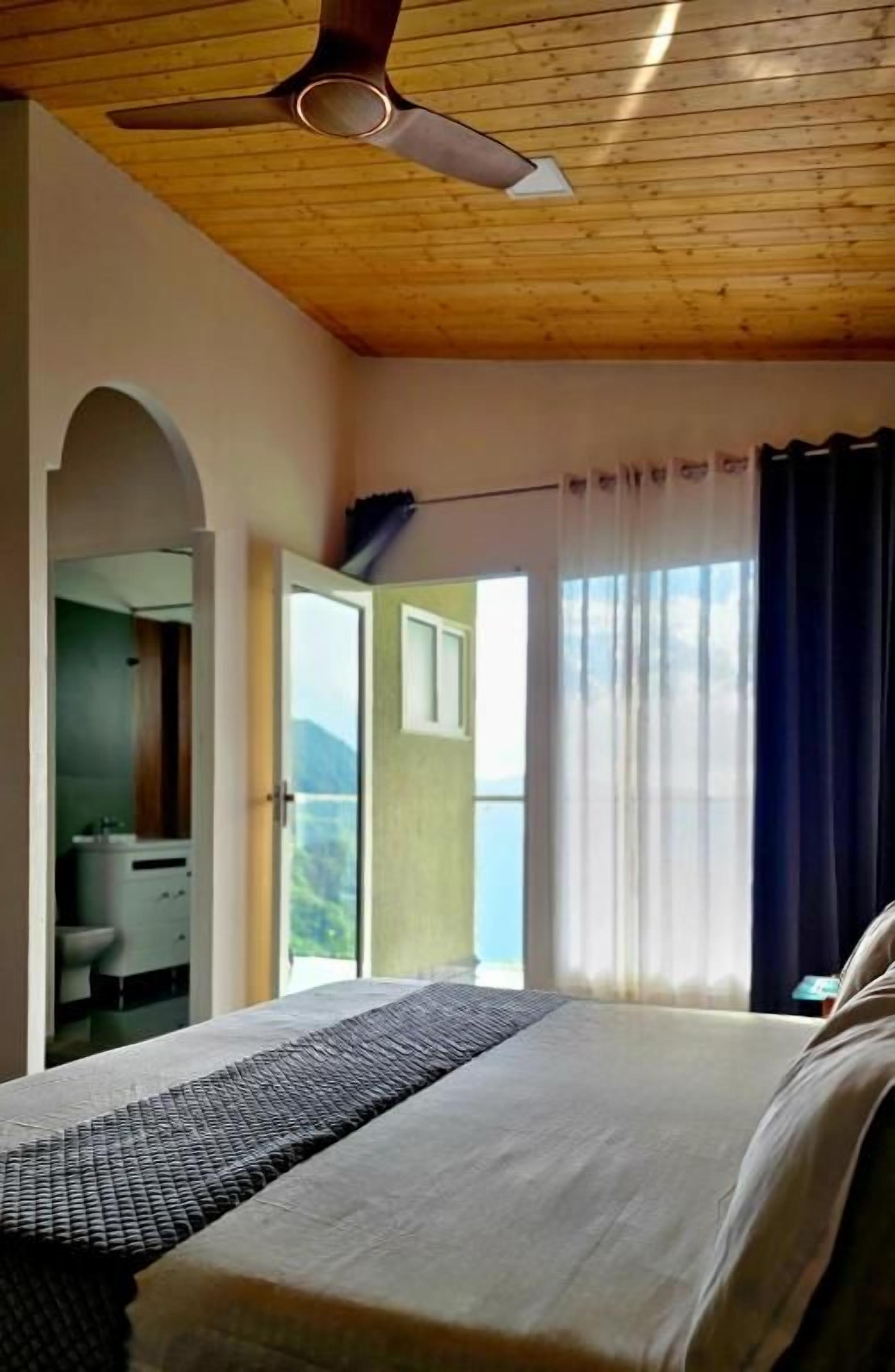 A bedroom with a bed, a ceiling fan, a window with curtains, and a view of the outdoors.