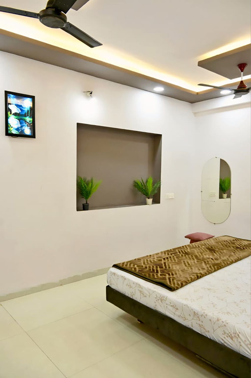 A bedroom with a bed, ceiling fan, recessed lighting, a wall-mounted mirror, and two potted plants.