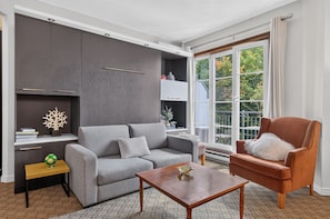 A modern living room with a grey sofa, a wooden coffee table, and a brown armchair. There is a bookshelf with decorative items and a window with a view of greenery. - Johannsen 359 (Mont-Tremblant)