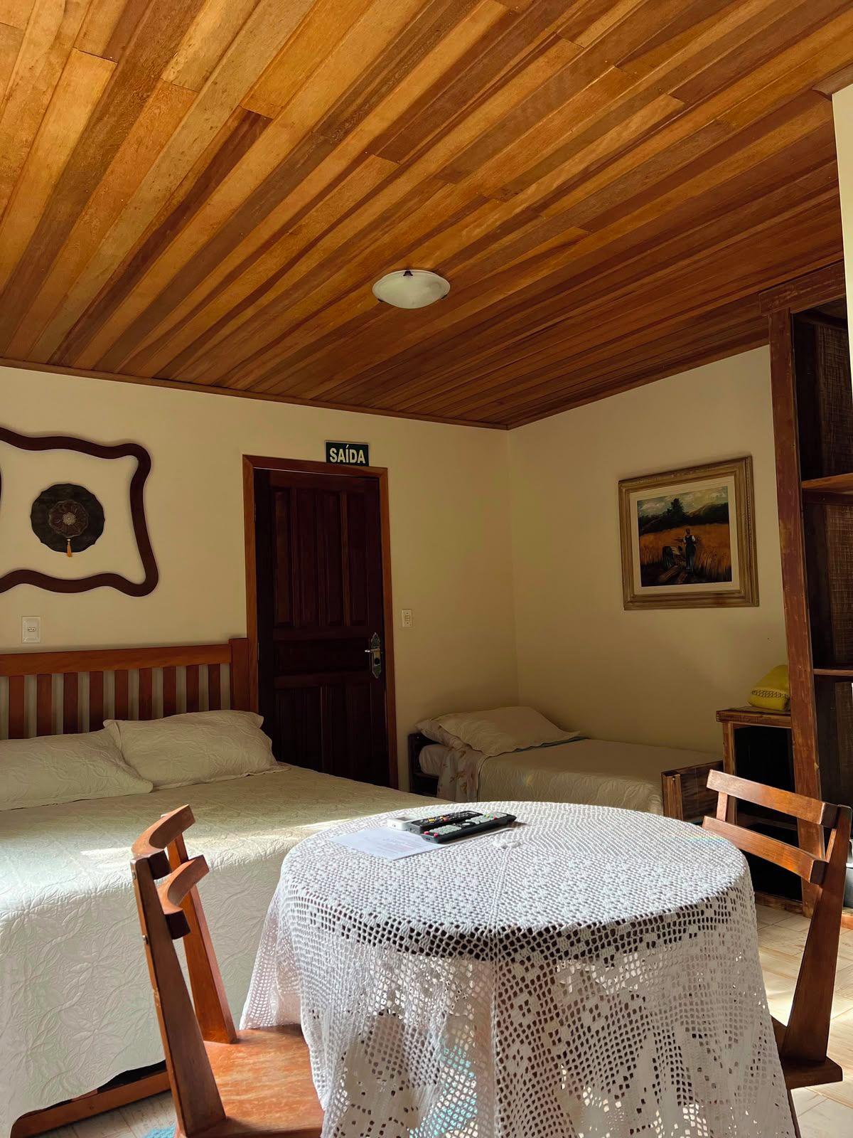 Een kamer met twee bedden, een houten plafond, een deur met de tekst 'SAÍDA', een schilderij aan de muur en een tafel met een kantkleed.