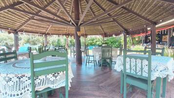 An outdoor covered patio with wooden tables and chairs, a thatched roof, and potted plants.