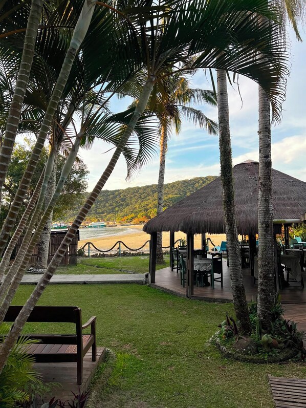 A thatched-roof outdoor seating area with tables and chairs, surrounded by palm trees and a view of a beach and hills.