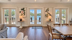A spacious room with a wooden floor, a dining table with chairs, and a view of a lake through large windows.