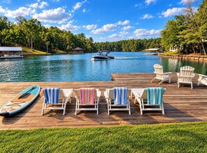 A wooden deck with lounge chairs and a paddleboard, overlooking a lake with a boat and dock.