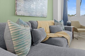 A modern living room with a grey sofa, multiple cushions, a throw blanket, and a large abstract painting on the wall. - Maré 2BR | Near Ocean Park Beach + Laundry (San Juan)