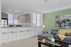 A modern kitchen with bar stools, a dining table, and a living area with a sofa and a painting. - Maré 2BR | Near Ocean Park Beach + Laundry (San Juan)