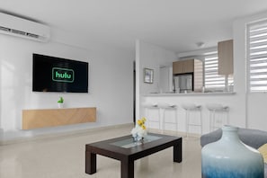 Una sala de estar moderna con un televisor de pantalla plana, una mesa de centro, un sofá y una cocina con taburetes de bar.