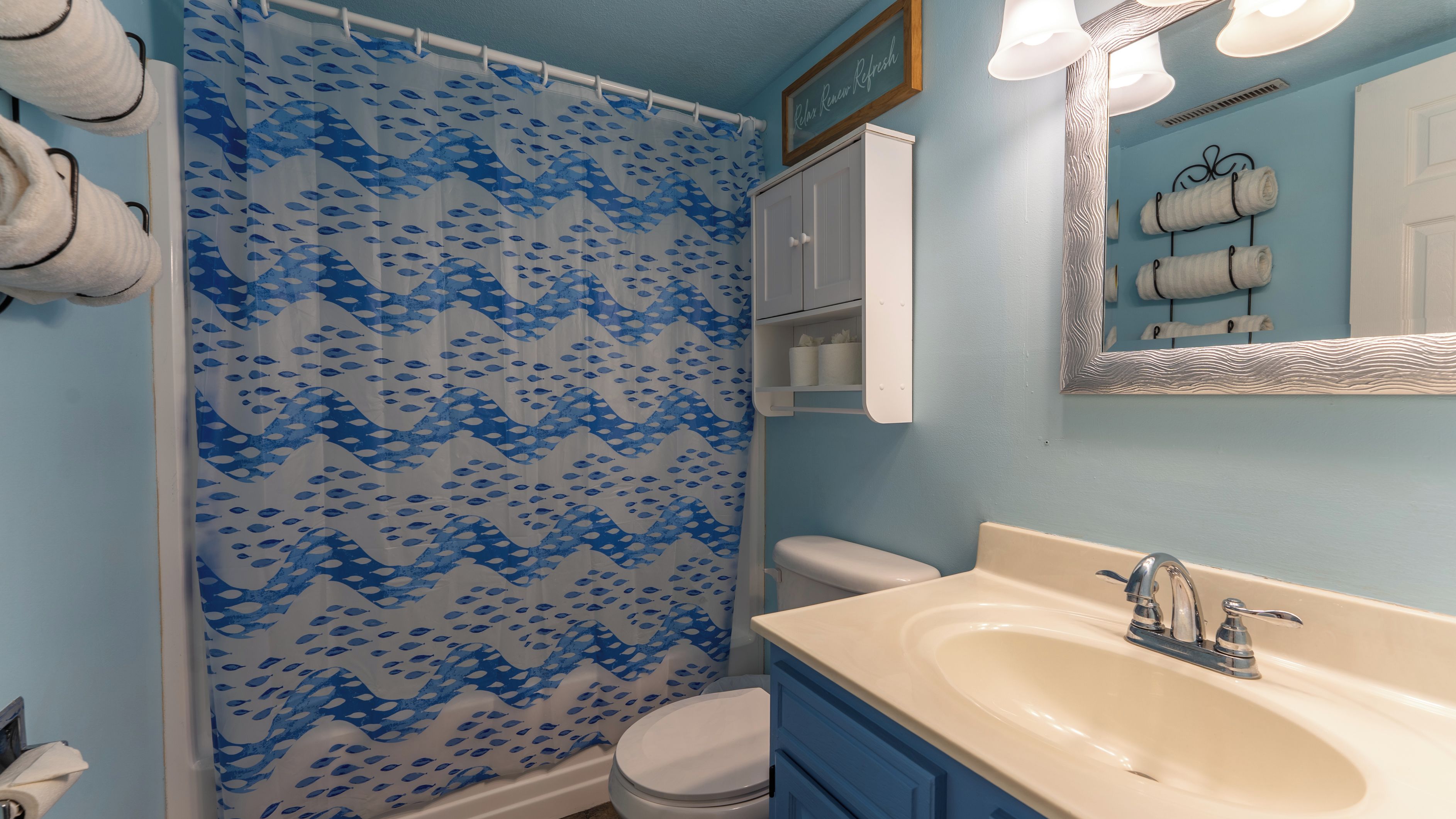 Un baño con cortina de ducha estampada con un motivo de peces azules, un inodoro blanco, un lavabo color beige con grifo plateado y un espejo con marco ondulado.