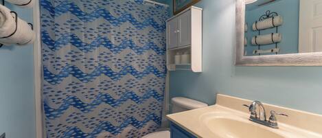 Un baño con cortina de ducha estampada con un motivo de peces azules, un inodoro blanco, un lavabo color beige con grifo plateado y un espejo con marco ondulado.