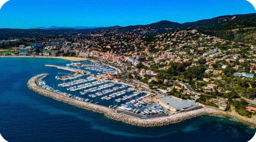 An aerial view of a coastal town with a marina, residential buildings, and a mountainous backdrop.