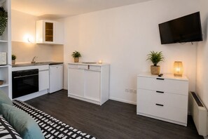 A compact kitchen with white cabinetry, a built-in oven, and a countertop with a sink. There is a flat-screen TV mounted on the wall, a potted plant on the counter, and a small cabinet with drawers.