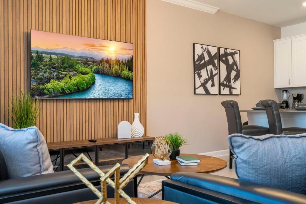 A modern living room with a flat-screen TV, two abstract paintings, a dining area with a round table, and a kitchen in the background.