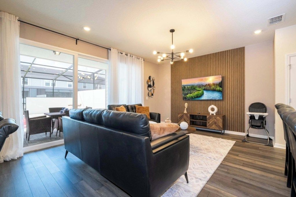 A modern living room with a black leather sofa, a flat-screen TV mounted on a wooden wall, a dining area with a table and chairs, and a large window with curtains.