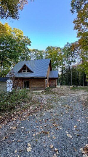 A wooden cabin with a metal roof, surrounded by trees and a gravel driveway. - Cozy Cabin Cottage on Big Gull Lake Waterfront (North Frontenac)