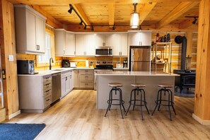 A modern kitchen with wooden cabinets, stainless steel appliances, and a central island with stools. - Cozy Cabin Cottage on Big Gull Lake Waterfront (North Frontenac)