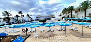 Uma área de piscina com vários espreguiçadeiras e guarda-sóis, cercada por palmeiras e edifícios.