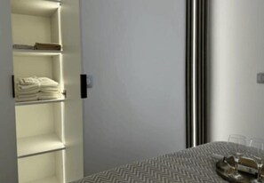 Quarto de hotel com uma cama, uma mesa posta com copos e um prato, e um guarda-roupa com toalhas dobradas.