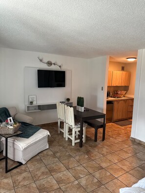 Un salon moderne comprenant une table à manger, des chaises, un téléviseur à écran plat fixé au mur, un canapé et une petite table d’appoint avec un panier.