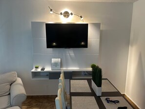 A modern living room with a flat-screen TV mounted on a white wall, a small plant on a shelf, and a couch. - Saint Tropez Great Apartment & Location (Carolina)