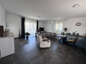 A modern living room with a sofa, a dining area with chairs, a kitchen area with a counter, and a large window with curtains. - Bravone House, 43 m² (linguizzetta)