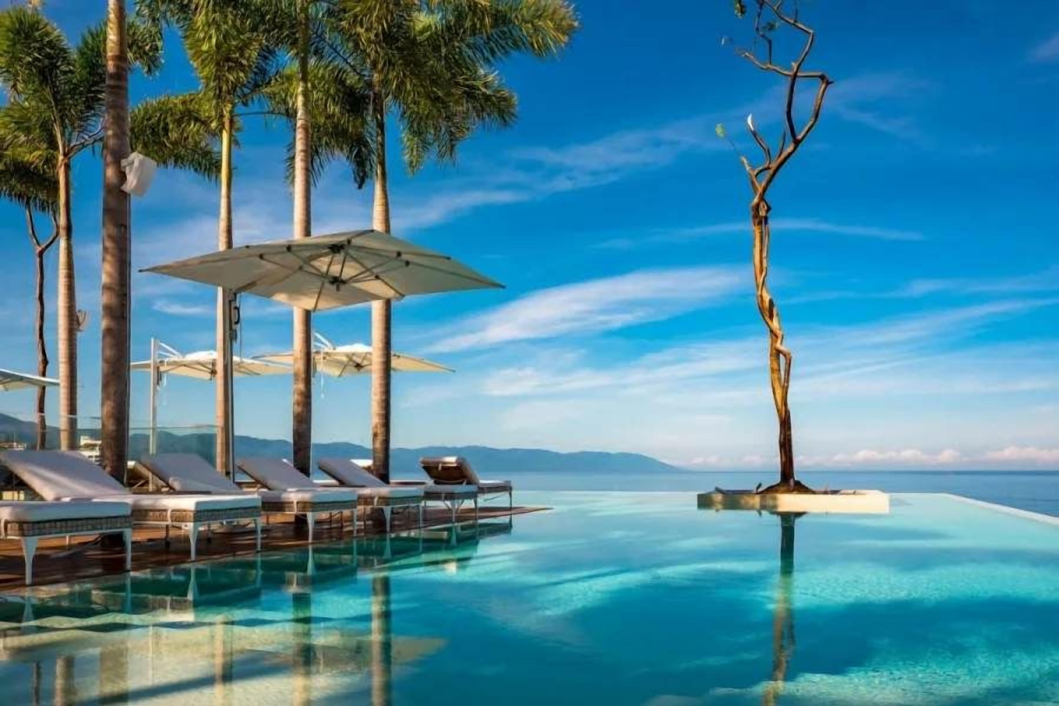 An infinity pool with lounge chairs and umbrellas, surrounded by palm trees and a clear sky.
