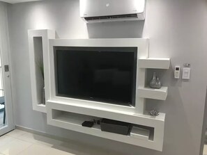 En moderne stue med et stort fladskærms-tv monteret på en hvid væg, en indbygget klimaanlæg ovenover, og en lille reol med pynteting.