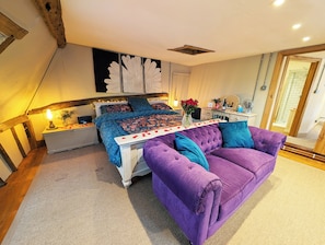 A bedroom with a bed, a purple sofa, a nightstand, a bedside table, and a wall-mounted decorative piece. - Chinnerys Country Cottage (Sudbury)