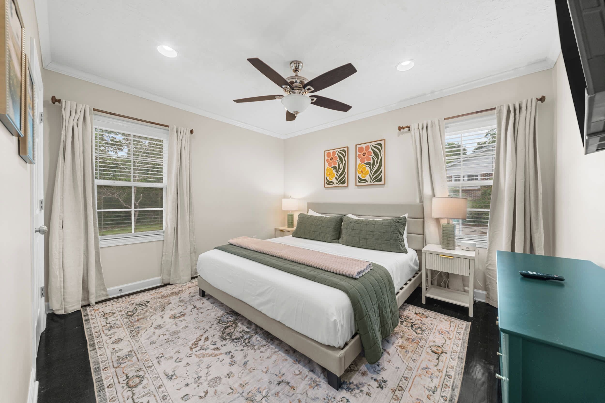 A bedroom with a bed, a ceiling fan, two wall-mounted lamps, a nightstand, a TV stand, and a window with curtains.
