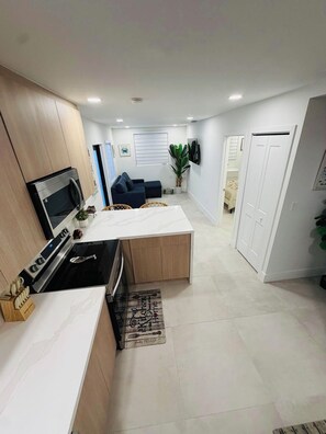 Eine moderne Küche mit integriertem Backofen und Mikrowelle, ein Essbereich mit weißem Tisch und ein Wohnzimmer mit Sofa und Fernseher.
