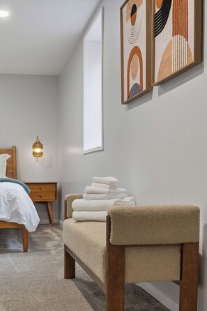 Ein Schlafzimmer mit einem Bett, einem Nachttisch, einer Lampe, einer Bank mit Handtüchern und abstrakter Wandkunst.