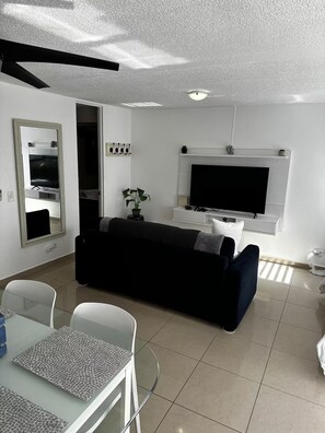 A modern living room with a black sofa, a flat-screen TV, a dining area with a white table, and a hallway with a mirror. - Best Location, Condado Studio, Beach Access (San Juan)