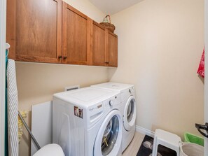 A laundry room with a front-loading washer and dryer, wooden cabinets above, and a small stool. - Quail Cottage with complimentary Golf Cart #187 (Osoyoos)