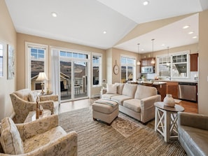 A spacious living room with a large window, a kitchen in the background, and a sliding glass door leading to a patio. - Quail Cottage with complimentary Golf Cart #187 (Osoyoos)