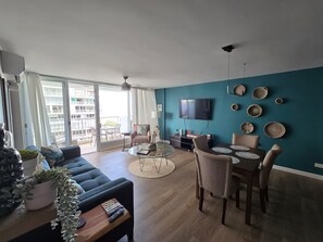 A modern living room with a sofa, coffee table, and dining area. A TV is mounted on the wall, and there are decorative items on the wall. - Spectacular Apt With Beach View (Carolina)