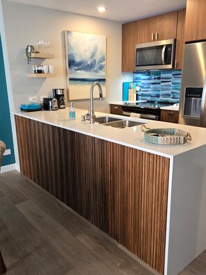 A modern kitchen with a central island, stainless steel appliances, and a coastal-themed artwork. - Spectacular Apt With Beach View (Carolina)