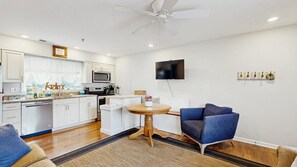 A modern kitchen with white cabinets, a microwave, and a dishwasher. There is a round wooden table with a white appliance on it. A blue armchair is visible in the foreground. - Beach Club - 20981 Coastal Hwy #4 (Rehoboth Beach)