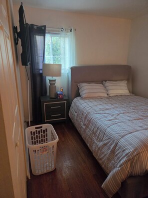 A bedroom with a bed, a nightstand, a lamp, a laundry basket, and a window with curtains. - Awesome 3-bedroom house with AC in Fort Washington near National Harbor & MGM (Fort Washington)