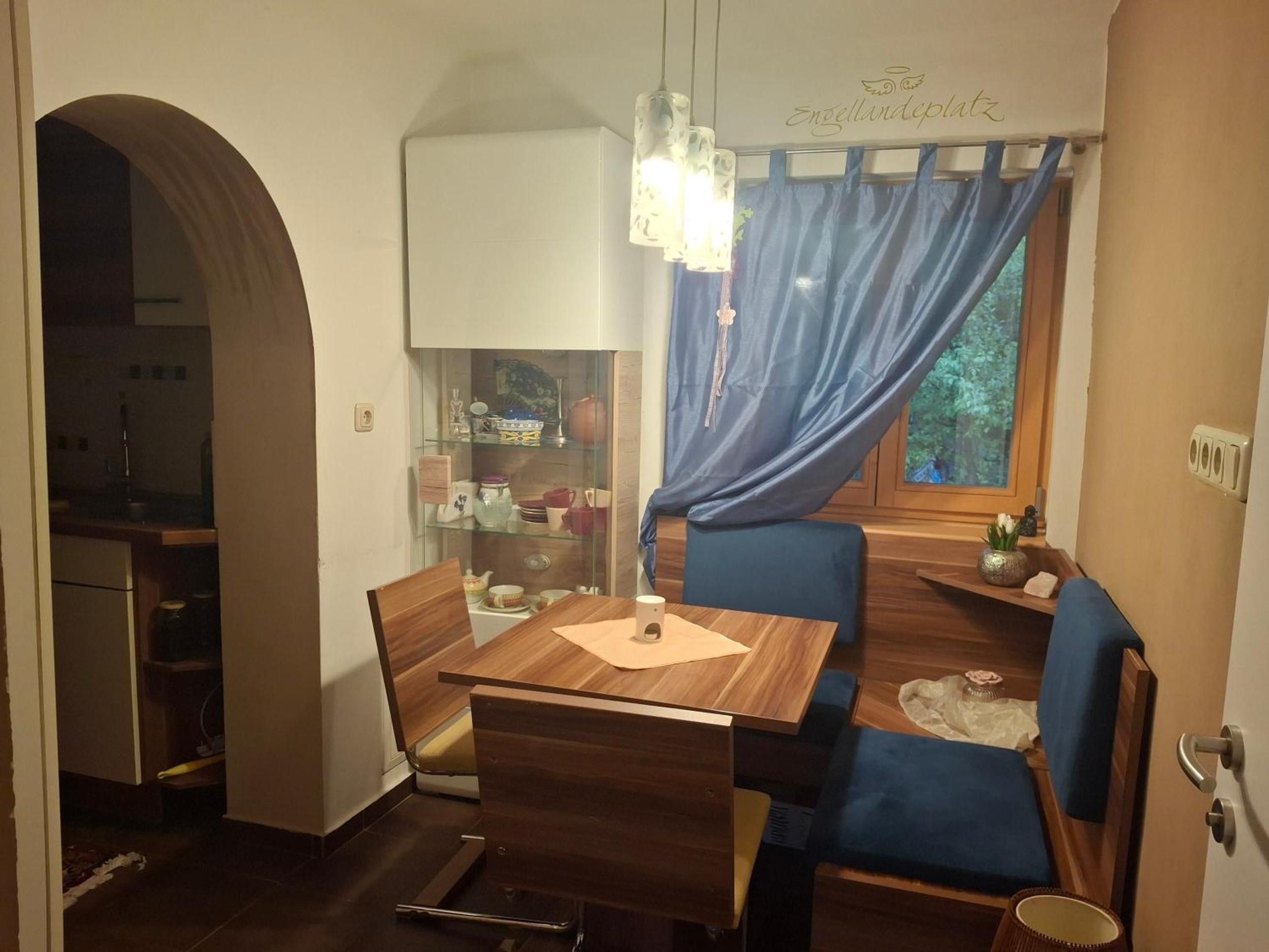 Uma área de refeições compacta com uma mesa e cadeiras de madeira, um armário de vidro com louças, uma cortina azul e uma janela com vista.