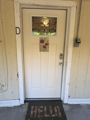 Een witte deur met een ruit, een deurmat waarop 'HELLO' staat en een klein bordje met rode bloemen.