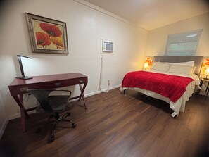 Een slaapkamer met een bed, een bureau met stoel, een wandairconditioner en een lijst met een afbeelding.