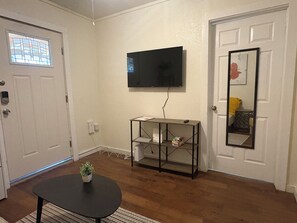 Een kamer met een wandgemonteerde flatscreen televisie, een kleine tafel met een bloempot en een deur met een raam.