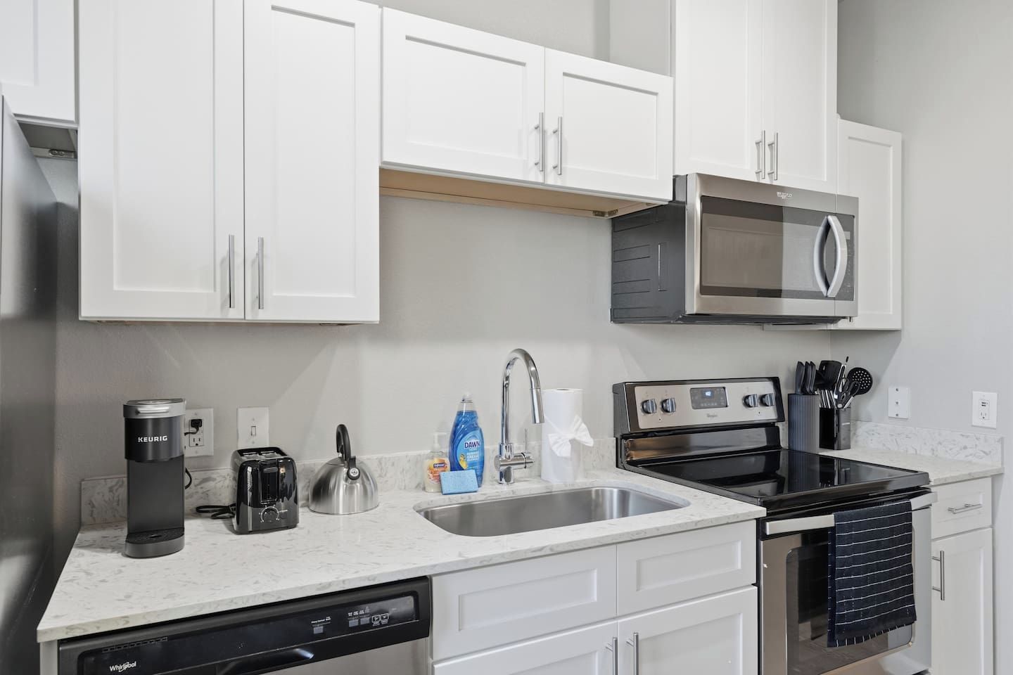 A modern kitchen with white cabinets, a stainless steel microwave, a coffee maker, a kettle, a dishwasher, and an oven.