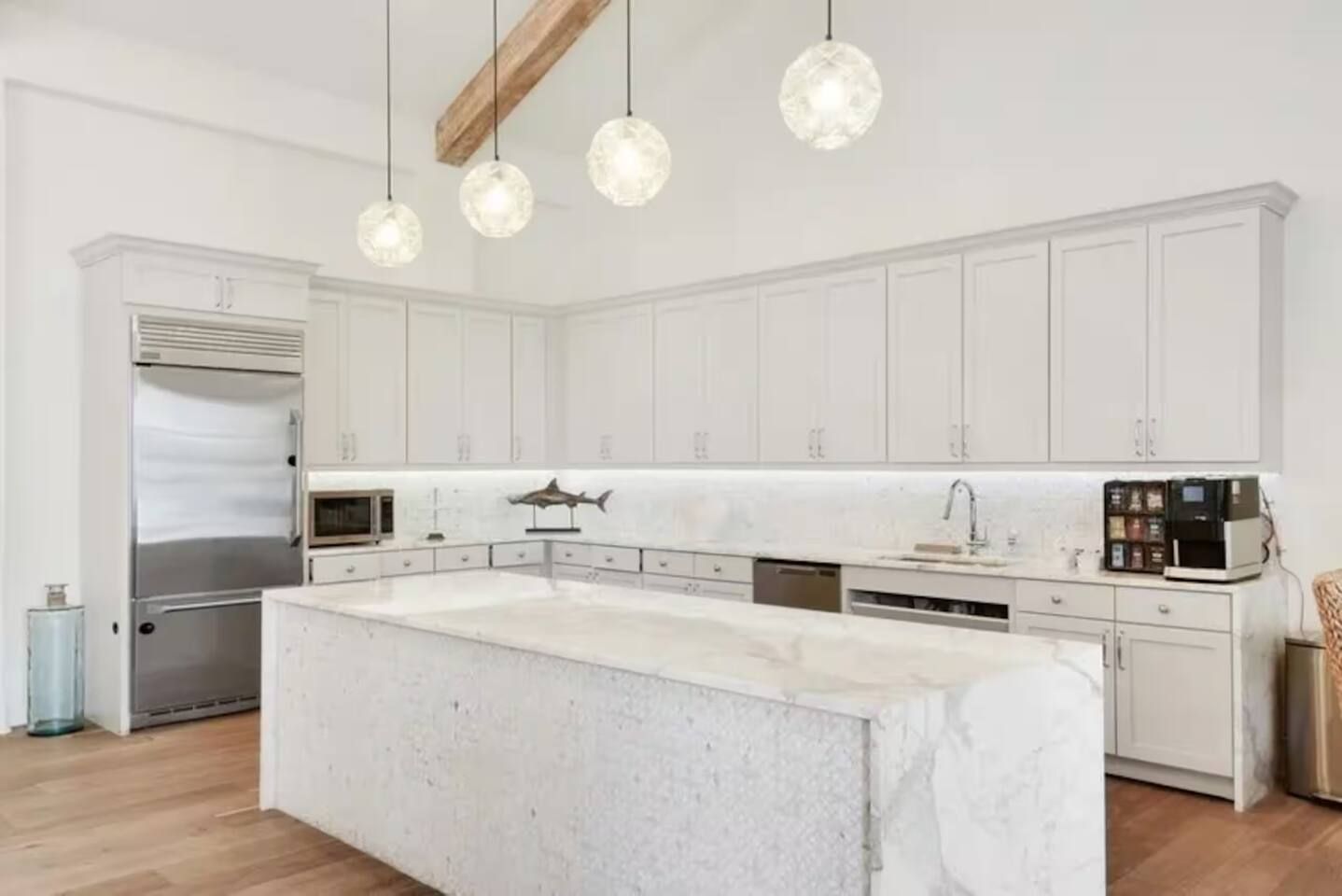 Une cuisine moderne avec des meubles blancs, un grand îlot central, des appareils en acier inoxydable et des luminaires suspendus.