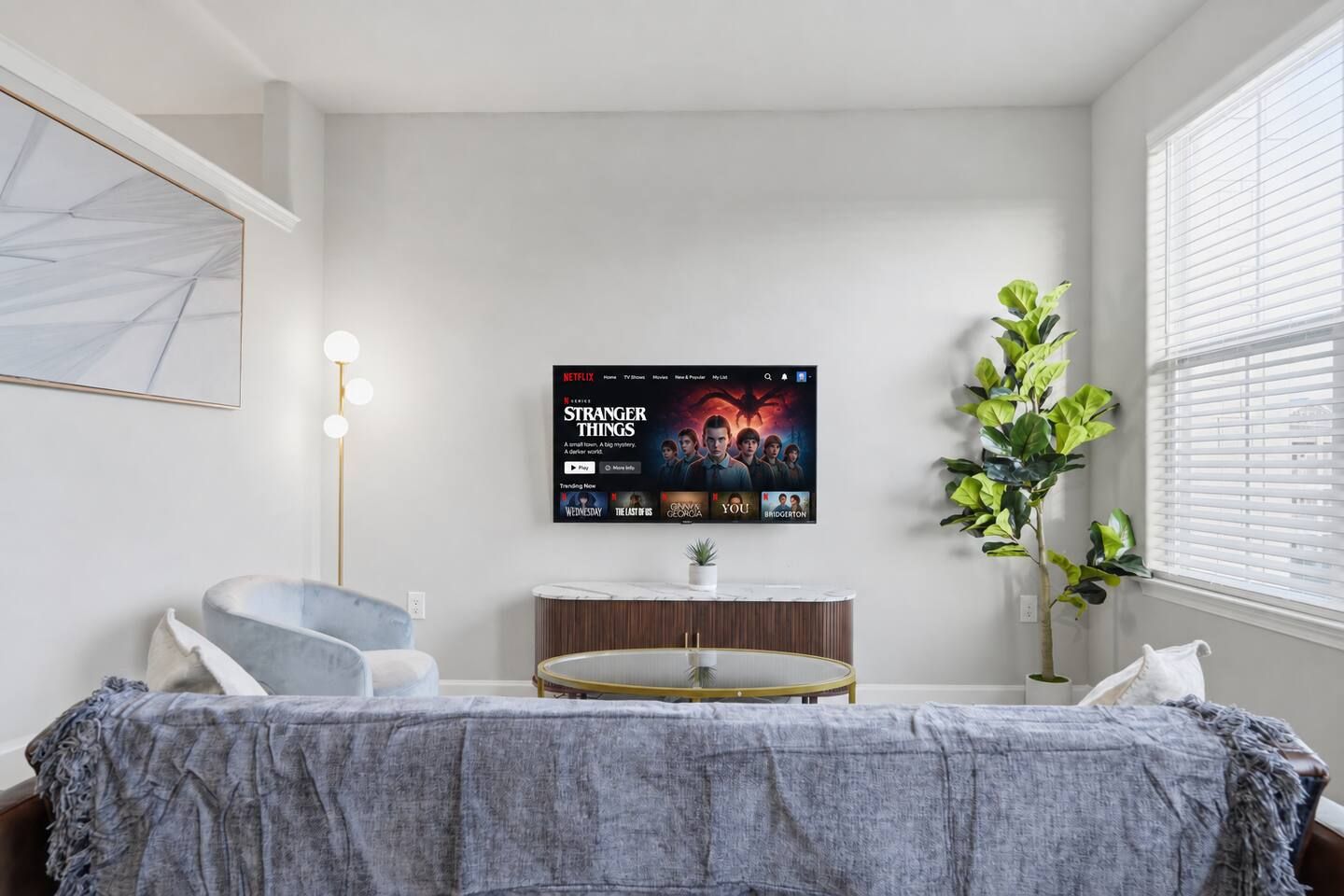 A modern living room with a flat-screen TV displaying 'Stranger Things', a grey sofa, a small round table with a plant, and a large window with blinds.