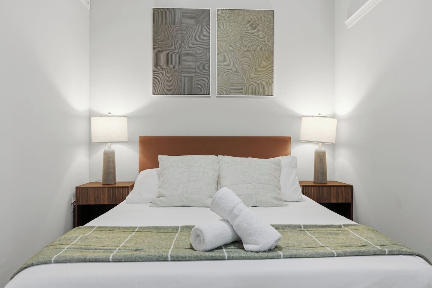 A hotel room with a bed, two bedside lamps, a headboard, and two wall art pieces.