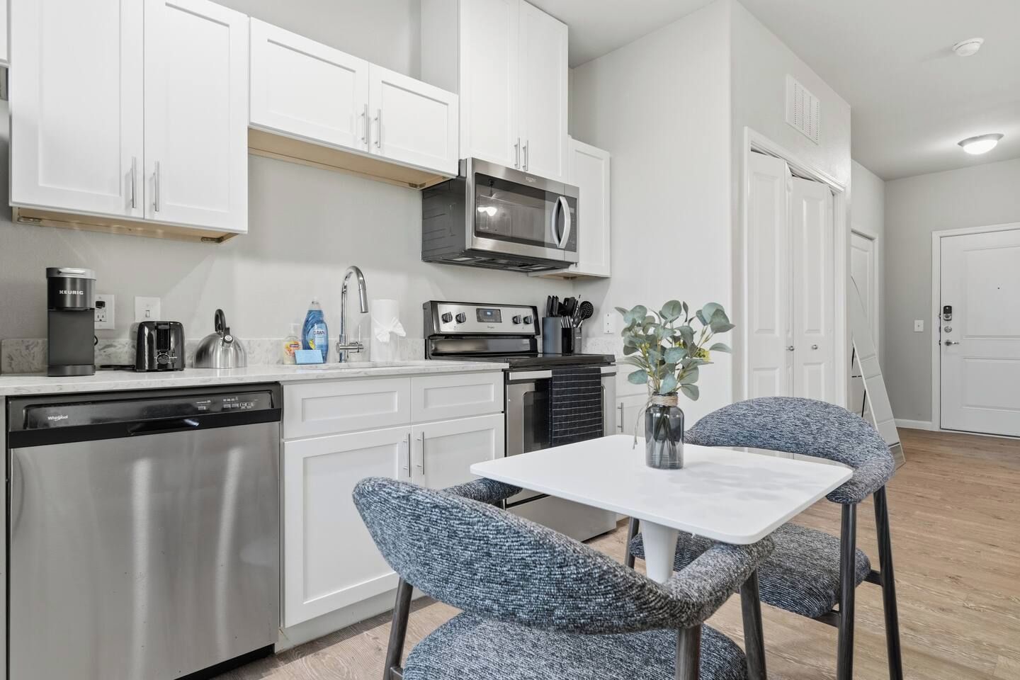 A modern kitchen with white cabinets, a built-in microwave, a dishwasher, and a small dining table with two chairs.