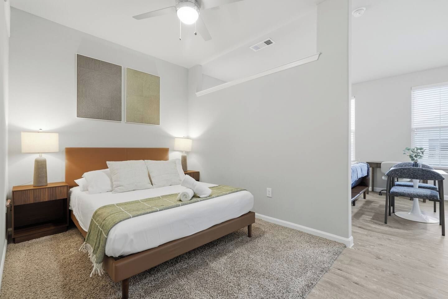 A hotel room with a bed, two bedside lamps, a ceiling fan, a small desk with a chair, and a window with blinds.