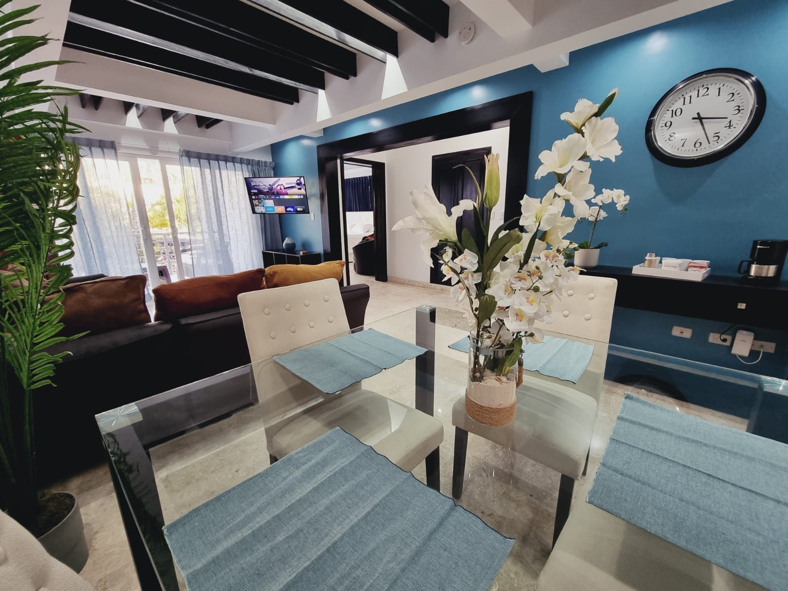 A modern living room with a glass dining table, a sofa, and a TV in the background.