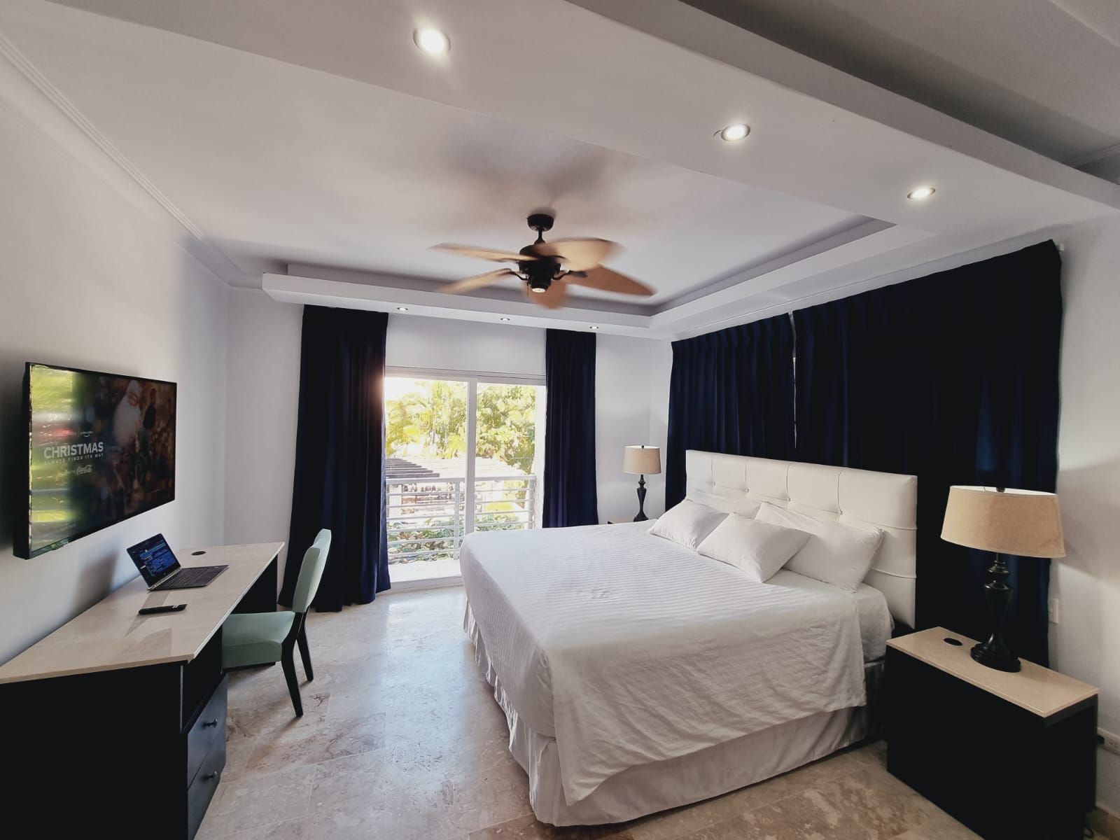 A hotel room with a bed, a desk with a laptop, a TV, and a ceiling fan.